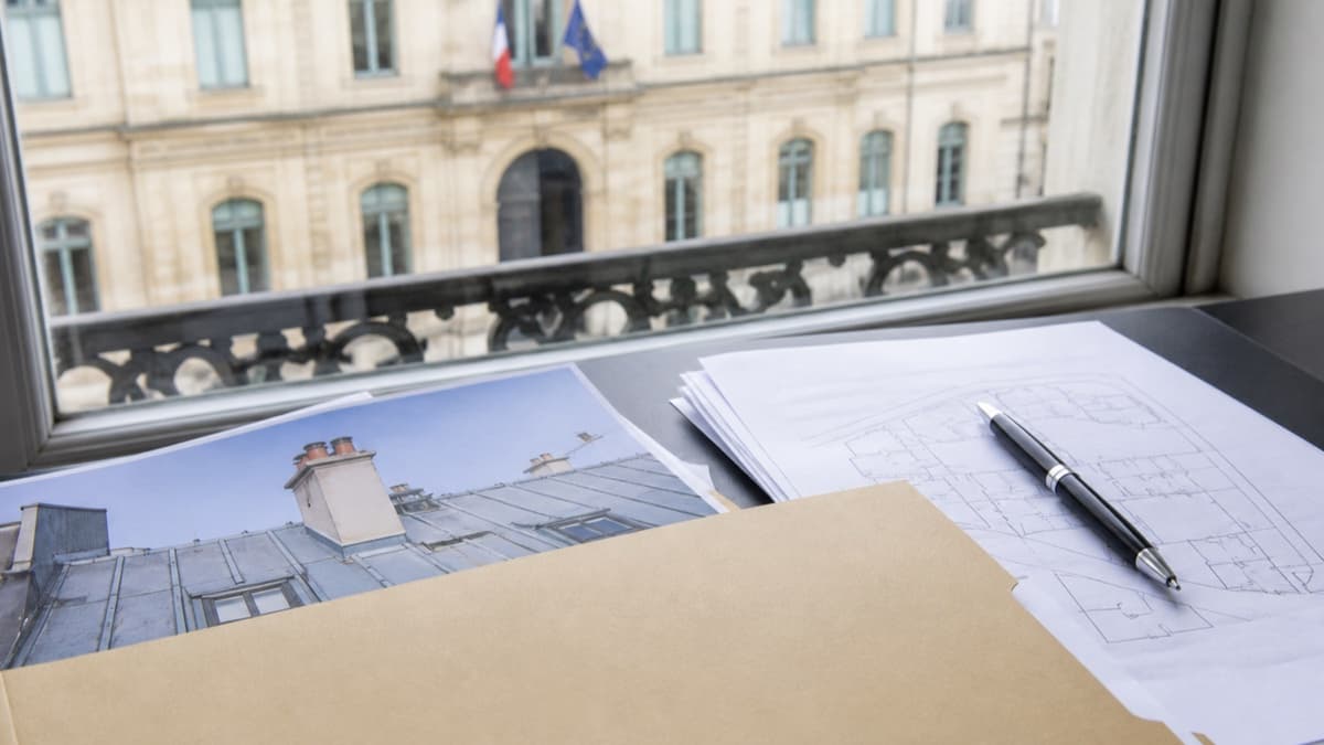 Dossier administratif de travaux toiture près d’une fenêtre donnant sur un bâtiment public